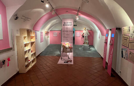 Das Bild zeigt einen Teil der Sonderaustellung Glück - Sonderausstellung Glück - Happiness im Museum der Stadt Villach. Das Bild zeigt eine gesamtübersicht über einen Raum der Sonderausstellung. Der Raum ist vorwiegend in weiß mit rosa Farbakzenten gehalten. Der Raum beschäftigt sich mit dem Theme Glück durch Konsum.
